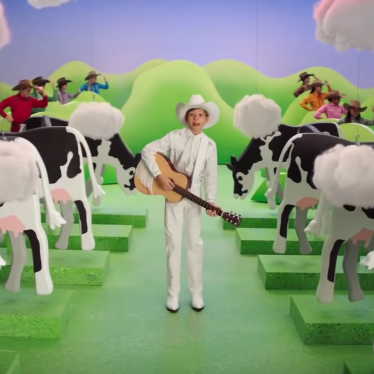 Video still of a boy with a guitar standing between two rows of cows.