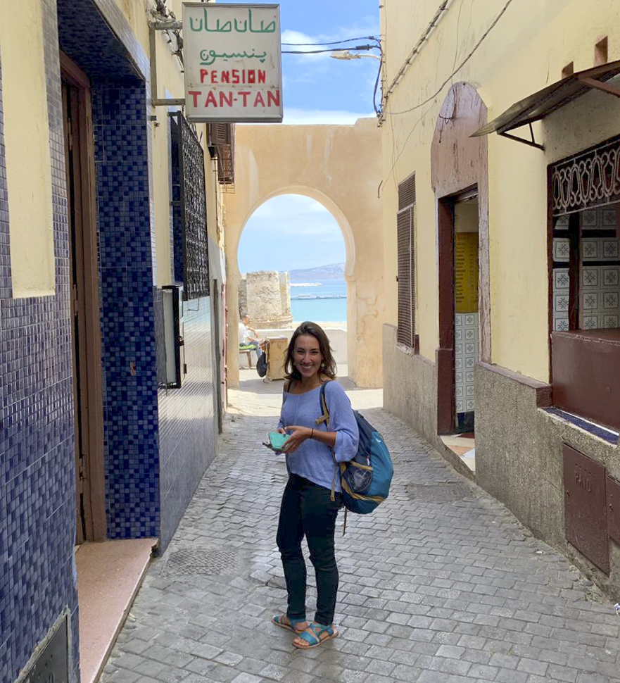 Lorena Gazzotti on fieldwork in Tangier. She is standing in an alleyway of a village with a view onto the sea.