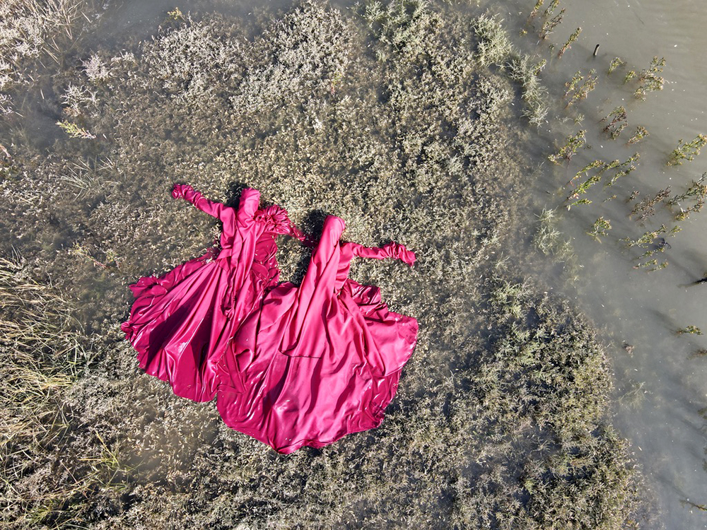 Two bright pink full body dresses laid out on a swampy shore.