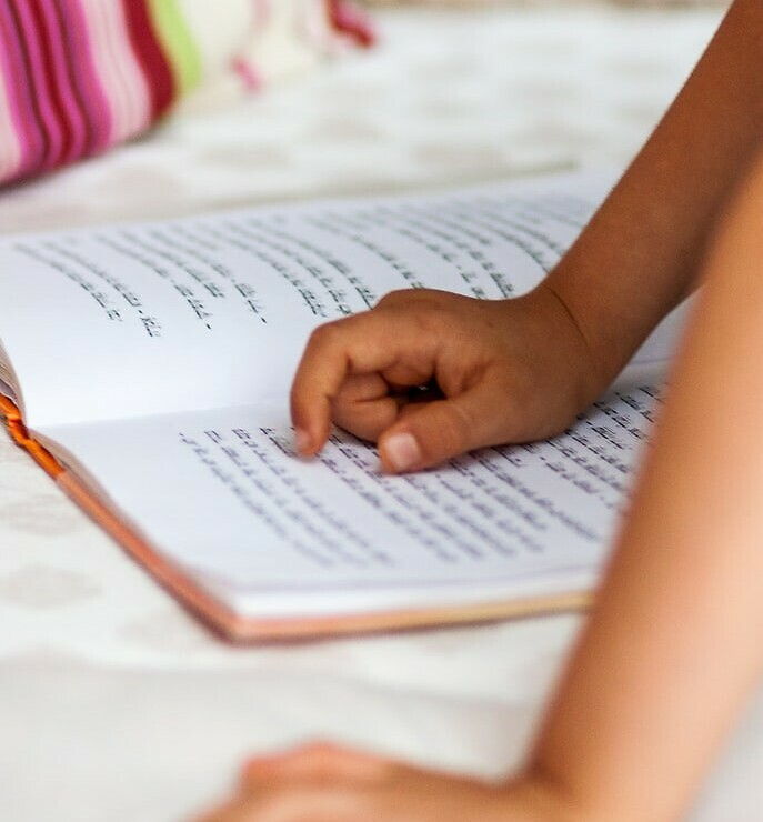 Photograph showing a child's hand, following the lines of text in a Hebrew book