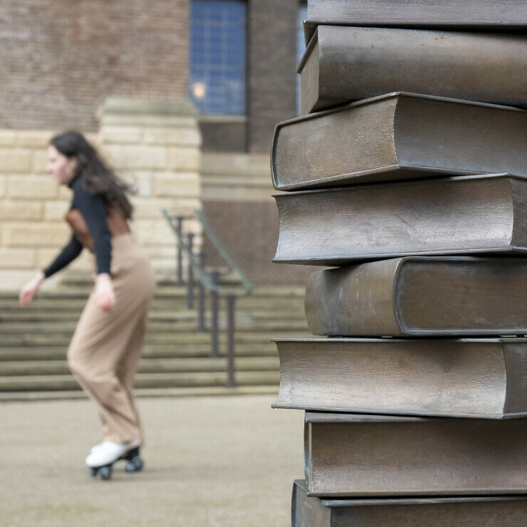 A woman rollerskates past a bronze bollard made from books in front of Cambridge University Library.