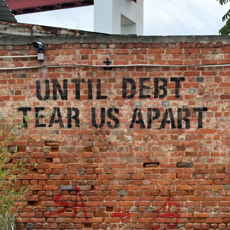 Backyard beneath a motorway bridge. At the back of the yard is a brick wall with graffiti: 'Until debt tear us apart'.