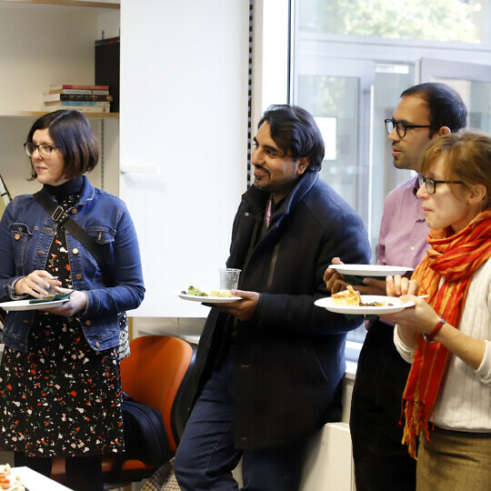 A group of fellows and researchers at the CRASSH welcome lunch. Everyone is holding plates of food and listening intently so someone who is speaking out of shot.