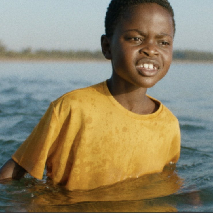 A young African boy with a yellow t-shirt is standing in water up to his chest.
