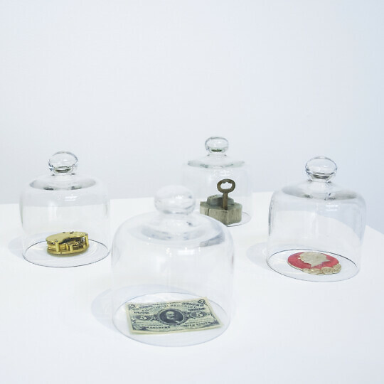 Various items are placed on a plinths under glass bell jars. Among others, they contain dollar bills and a tin of food with a tin opener attached.