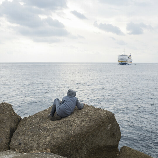Person sitting on a rock looking out to sea at a large ship.